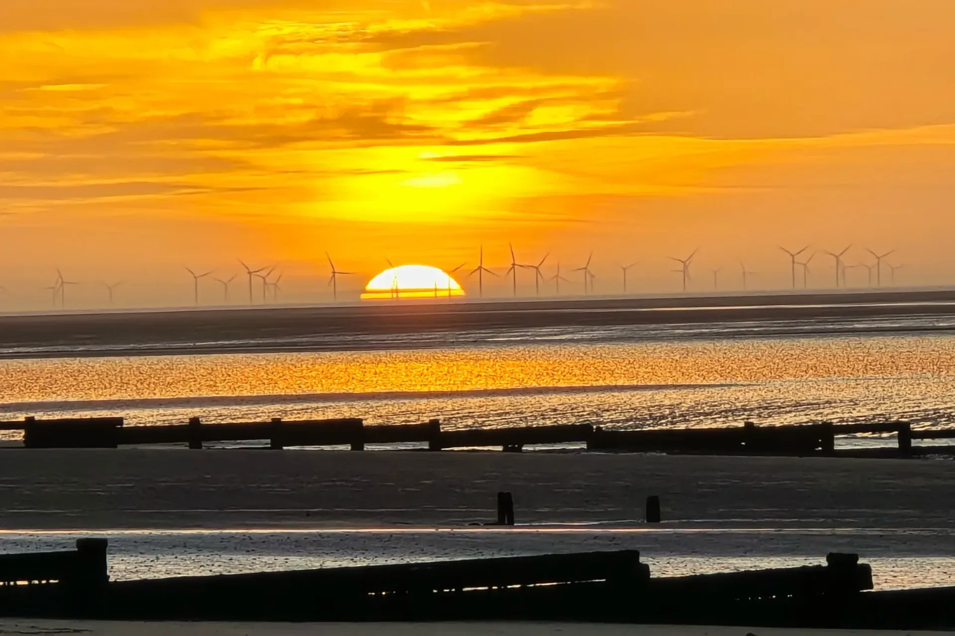 The sun sets into the sea behind a long line of wind turbines. Wooden groynes cross the wet sand in front.